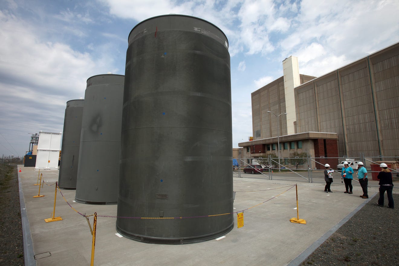 alt="Dry casks holding spent fuel assemblies are shown outside the Pilgrim Nuclear Power Station before its May 2019 shutdown. Owner Holtec International has reached an agreement with the state to ensure safe decommissioning of the plant and cleanup of the site."